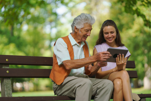 A young caregiver spending quality time with lonely senior client in the city park. The grandfather and granddaughter are looking at photos on a tablet, sitting on a bench in a city park.