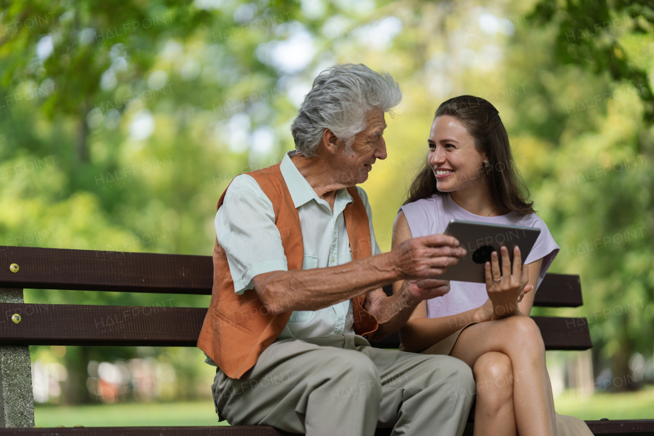 A young caregiver spending quality time with lonely senior client in the city park. The grandfather and granddaughter are looking at photos on a tablet, sitting on a bench in a city park.