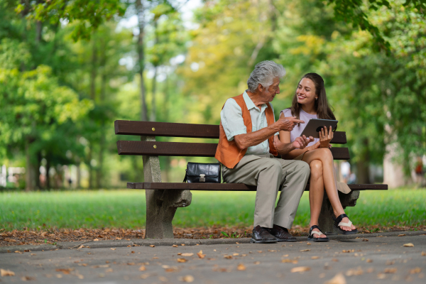 A young caregiver spending quality time with lonely senior client in the city park. The grandfather and granddaughter are looking at photos on a tablet, sitting on a bench in a city park.