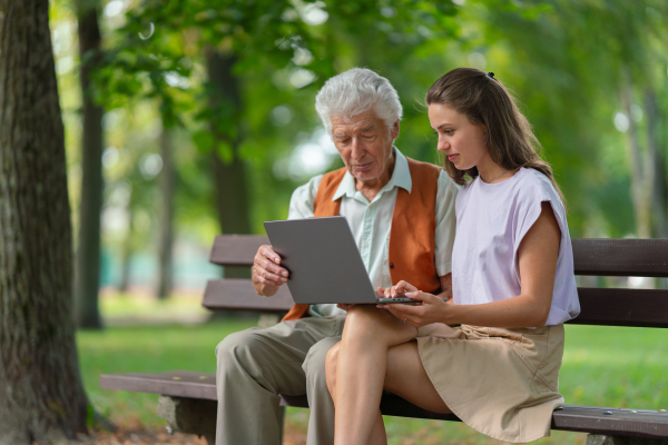 A young caregiver spending quality time with lonely senior client in the city park. The grandfather and granddaughter are looking at photos on a notebook, shopping online together.