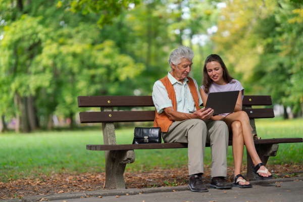 A young caregiver spending quality time with lonely senior client in the city park. The grandfather and granddaughter are looking at photos on a tablet, sitting on a bench in a city park.