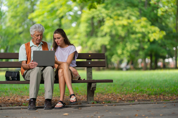 A young caregiver spending quality time with lonely senior client in the city park. The grandfather and granddaughter are looking at photos on a notebook, shopping online together.