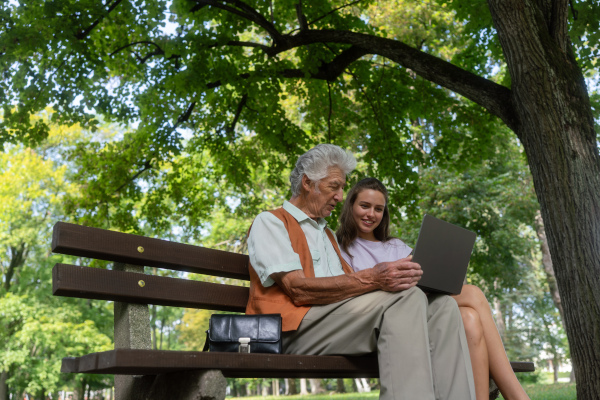 A young caregiver spending quality time with lonely senior client in the city park. The grandfather and granddaughter are looking at photos on notebook sitting on a bench in a city park. Young assistant teaching elderly man how to work with notebook, how to write email and work with internetbanking.