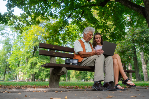 A young caregiver spending quality time with lonely senior client in the city park. The grandfather and granddaughter are looking at photos on notebook sitting on a bench in a city park. Young assistant teaching elderly man how to work with notebook, how to write email and work with internetbanking.