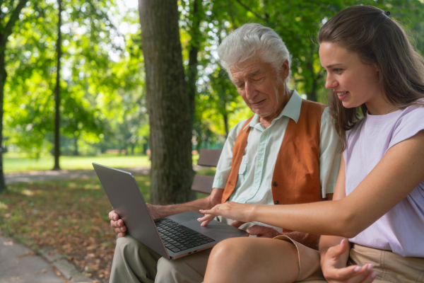 A young caregiver spending quality time with lonely senior client in the city park. The grandfather and granddaughter are looking at photos on notebook sitting on a bench in a city park. Young assistant teaching elderly man how to work with notebook, how to write email and work with internetbanking.