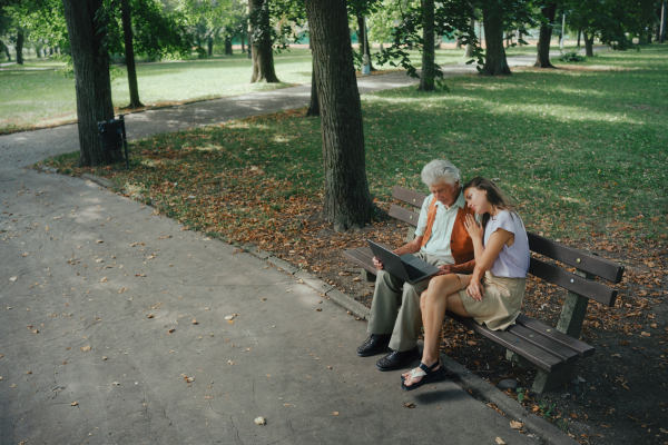 A widower is sitting on a bench in the park with his granddaughter, mourning his deceased wife. Looking at photos of grandma on laptop, reminiscing, feeling grief and sorrow.