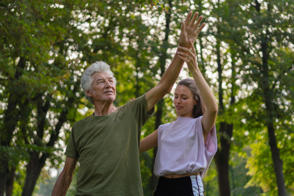 The senior man exercising in the park, A young caregiver showing older man how to perform the exercise correctly. A portrait of a young physiotherapist training outdoors in the park with senior patient.