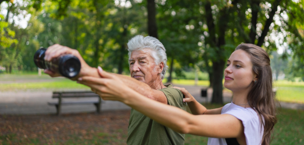 The senior man exercising in the park, using a water bottle as a dumbbell. A young caregiver showing older man how to perform the exercise correctly. A portrait of a young physiotherapist training outdoors in the park with senior patient. Banner with copy space.