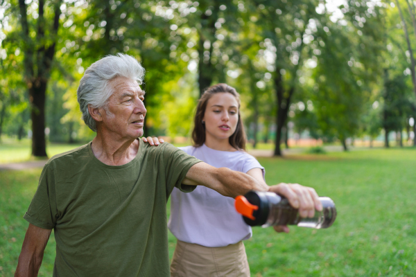 The senior man exercising in the park, using a water bottle as a dumbbell. A young caregiver showing older man how to perform the exercise correctly. A portrait of a young physiotherapist training outdoors in the park with senior patient. .