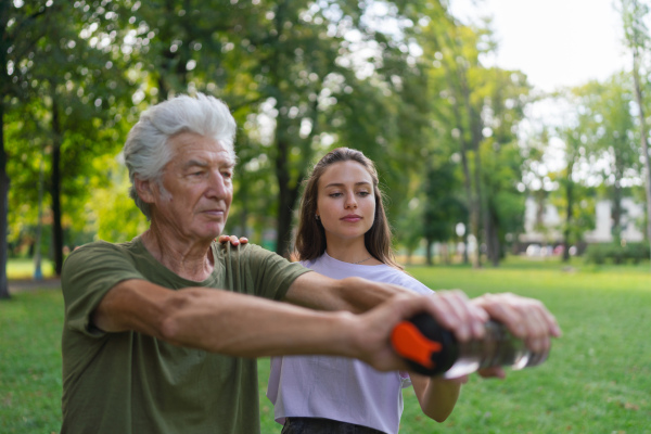 The senior man exercising in the park, using a water bottle as a dumbbell. A young caregiver showing older man how to perform the exercise correctly. A portrait of a young physiotherapist training outdoors in the park with senior patient. .