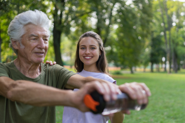 The senior man exercising in the park, using a water bottle as a dumbbell. A young caregiver showing older man how to perform the exercise correctly. A portrait of a young physiotherapist training outdoors in the park with senior patient. .