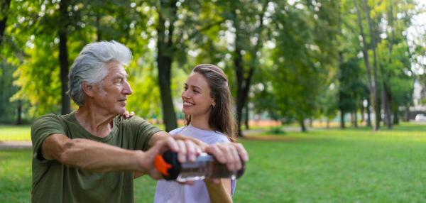 The senior man exercising in the park, using a water bottle as a dumbbell. A young caregiver showing older man how to perform the exercise correctly. A portrait of a young physiotherapist training outdoors in the park with senior patient. Banner with copy space.