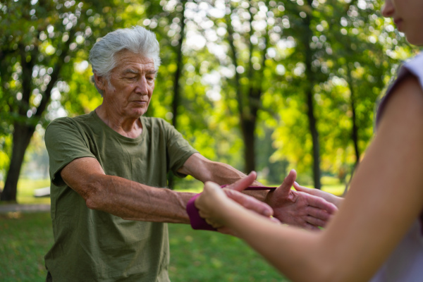 The senior man exercising in the park, using resistance band. A young caregiver showing older man how to perform the exercise with resistance band correctly. A portrait of a young physiotherapist training outdoors in the park with senior patient. .