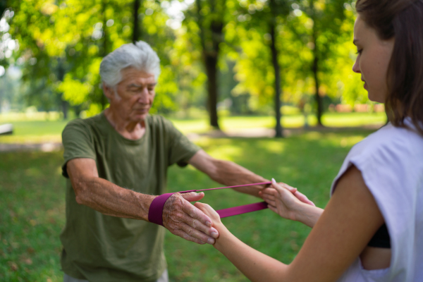 The senior man exercising in the park, using resistance band. A young caregiver showing older man how to perform the exercise with resistance band correctly. A portrait of a young physiotherapist training outdoors in the park with senior patient. .