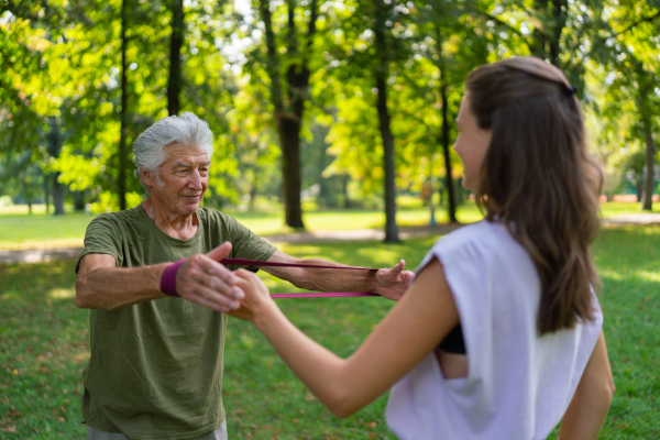 The senior man exercising in the park, using resistance band. A young caregiver showing older man how to perform the exercise with resistance band correctly. A portrait of a young physiotherapist training outdoors in the park with senior patient. .