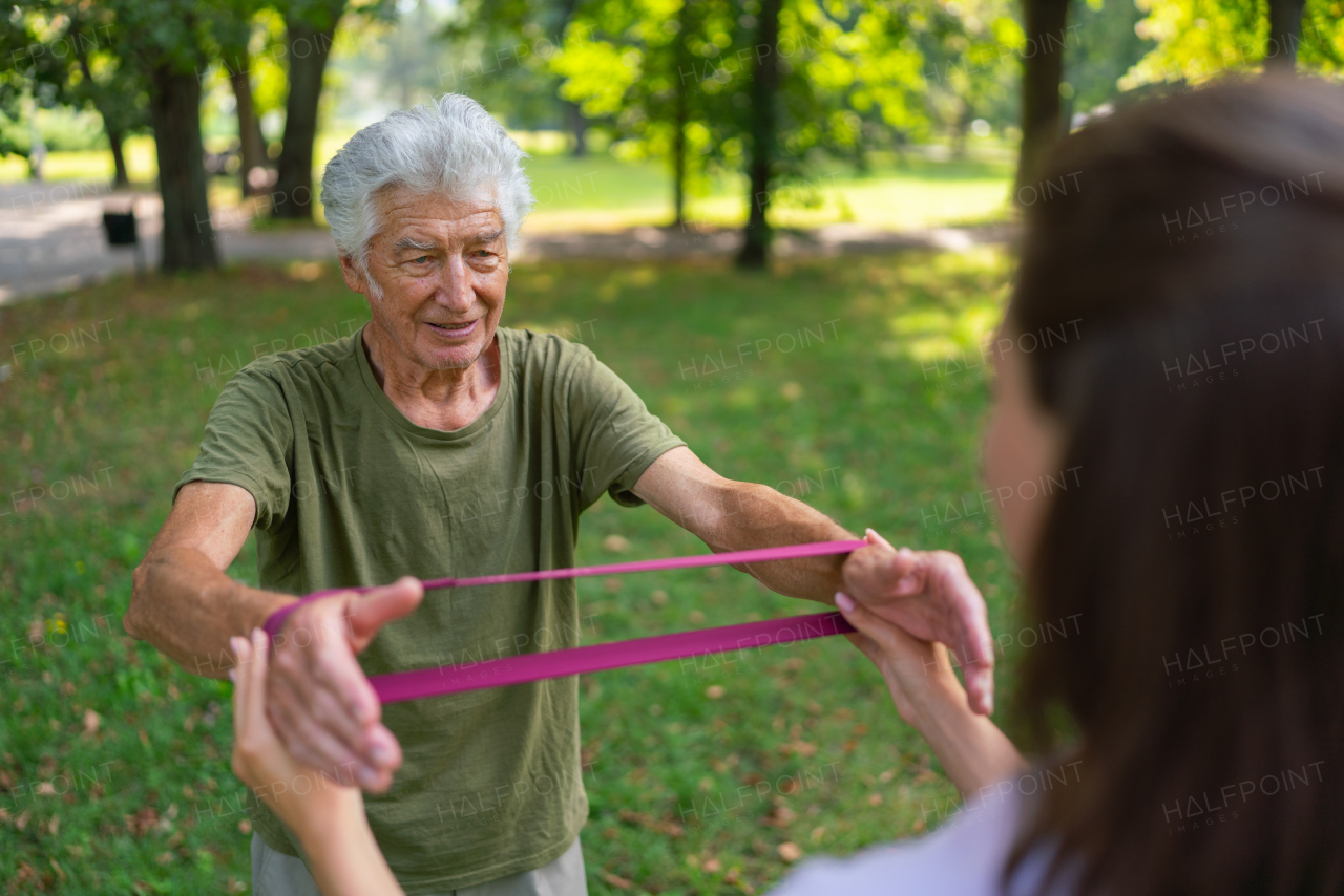 The senior man exercising in the park, using resistance band. A young caregiver showing older man how to perform the exercise with resistance band correctly. A portrait of a young physiotherapist training outdoors in the park with senior patient. .