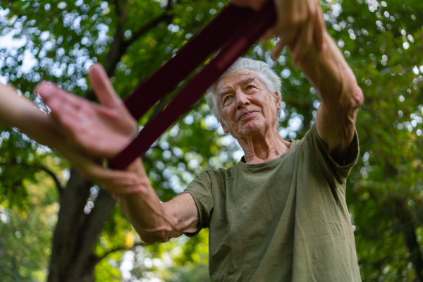 The senior man exercising in the park, using resistance band. A young caregiver showing older man how to perform the exercise with resistance band correctly. A portrait of a young physiotherapist training outdoors in the park with senior patient. .