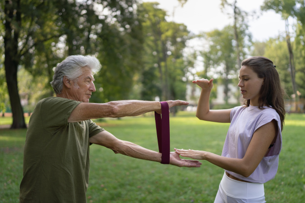 The senior man exercising in the park, using resistance band. A young caregiver showing older man how to perform the exercise with resistance band correctly. A portrait of a young physiotherapist training outdoors in the park with senior patient. .