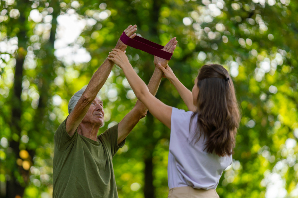 The senior man exercising in the park, using resistance band. A young caregiver showing older man how to perform the exercise with resistance band correctly. A portrait of a young physiotherapist training outdoors in the park with senior patient. .