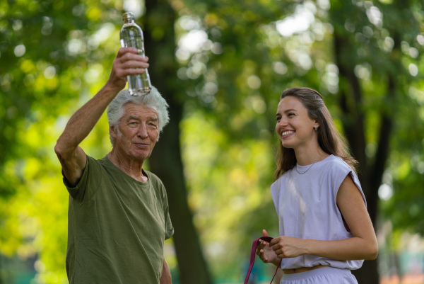 The senior man drinking water during workout, staying hydrated. Young physiotherapist training outdoors in the park with senior patient, letting him rest after exercising.