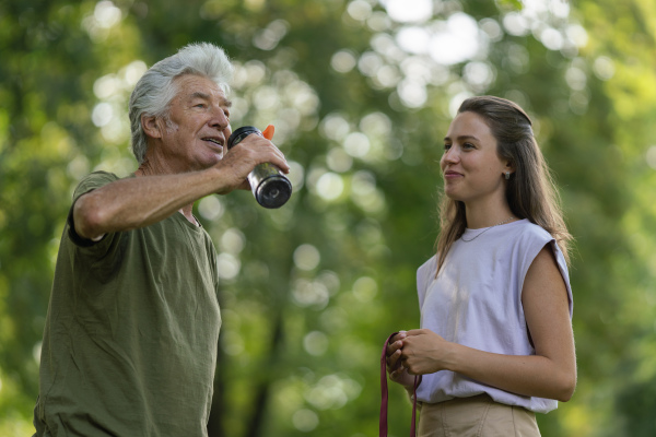 The senior man drinking water during workout, staying hydrated. Young physiotherapist training outdoors in the park with senior patient, letting him rest after exercising.