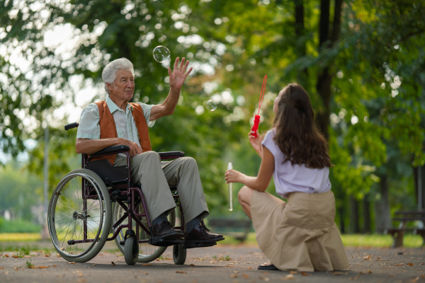 The man in a wheelchair having fun with his caregiver in the park, catching bubbles. The senior man in the wheelchair performs simple exercises, moving as he catches bubbles blown by the young nurse.