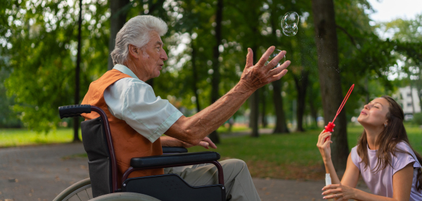 The man in a wheelchair having fun with his caregiver in the park, catching bubbles. The senior man in the wheelchair performs simple exercises, moving as he catches bubbles blown by the young nurse. Banner with copy space.