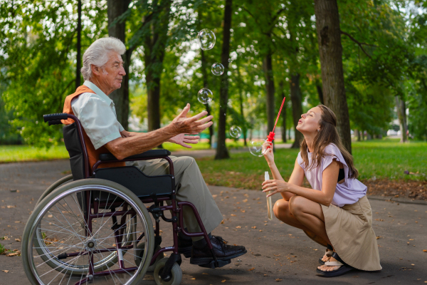 The man in a wheelchair having fun with his caregiver in the park, catching bubbles. The senior man in the wheelchair performs simple exercises, moving as he catches bubbles blown by the young nurse.