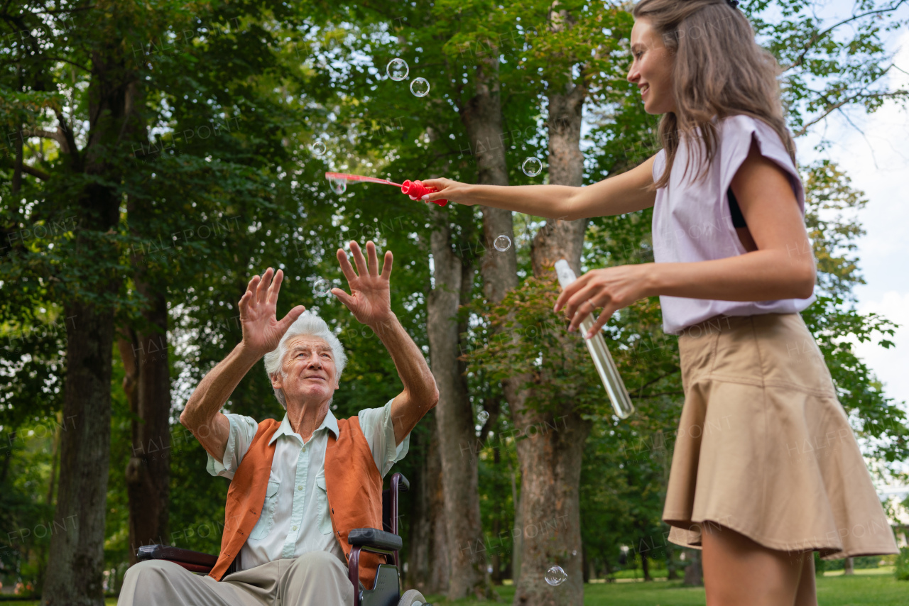 The man in a wheelchair having fun with his caregiver in the park, catching bubbles. The senior man in the wheelchair performs simple exercises, moving as he catches bubbles blown by the young nurse.