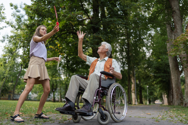 The man in a wheelchair having fun with his caregiver in the park, catching bubbles. The senior man in the wheelchair performs simple exercises, moving as he catches bubbles blown by the young nurse.