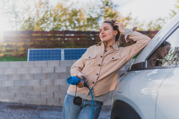 Young woman holding power supply cable from her car, prepared for charging it in home, sustainable and economic transportation concept.