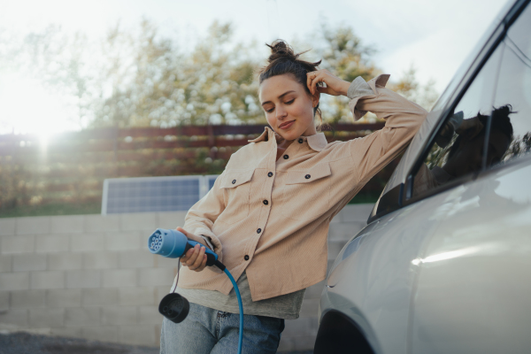 Young woman holding power supply cable from her car, prepared for charging it in home, sustainable and economic transportation concept.