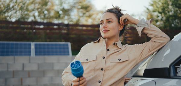 Young woman holding power supply cable from her car, prepared for charging it in home, sustainable and economic transportation concept.