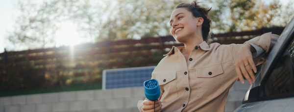 Young woman holding power supply cable from her car, prepared for charging it in home, sustainable and economic transportation concept.