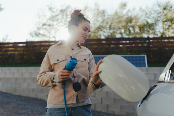 Young woman holding power supply cable from her car, prepared for charging it in home, sustainable and economic transportation concept.