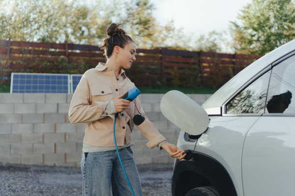 Young woman holding power supply cable from her car, prepared for charging it in home, sustainable and economic transportation concept.