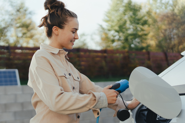 Young woman holding power supply cable from her car, prepared for charging it in home, sustainable and economic transportation concept.