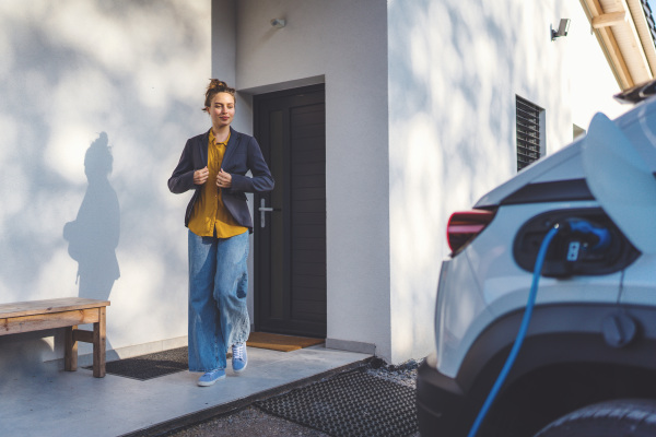 Young woman preparing to work while her electric car charging in own charging station in home, sustainable and economic transportation concept.