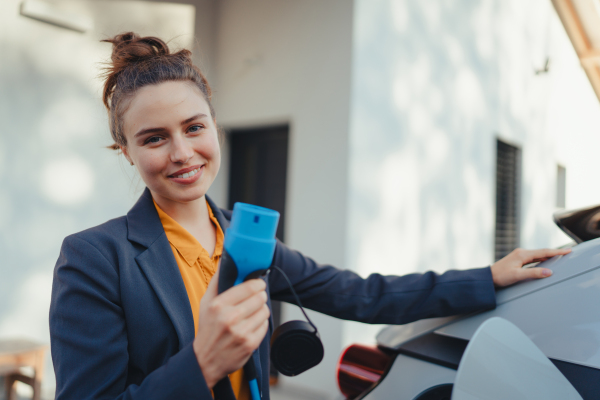 Young woman holding power supply cable from her car, prepared for charging it in home, sustainable and economic transportation concept.