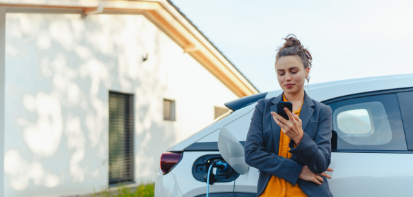 Young woman with smartphone waiting while electric car charging in home charging station, sustainable and economic transportation concept.