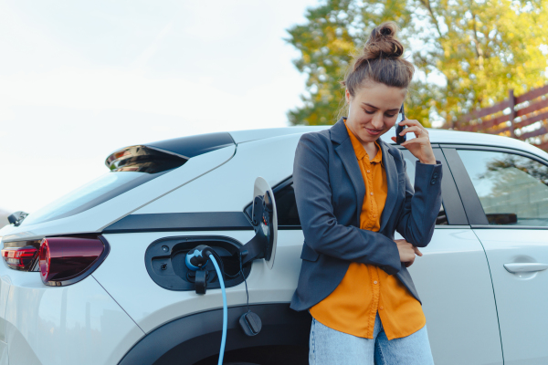 Young woman with smartphone waiting while electric car charging in home charging station, sustainable and economic transportation concept.