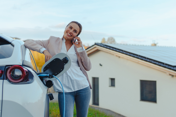 Young woman with smartphone waiting while electric car charging in home charging station, sustainable and economic transportation concept.