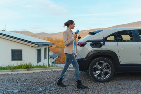 Young woman holding power supply cable from her car, prepared for charging it in home, sustainable and economic transportation concept.