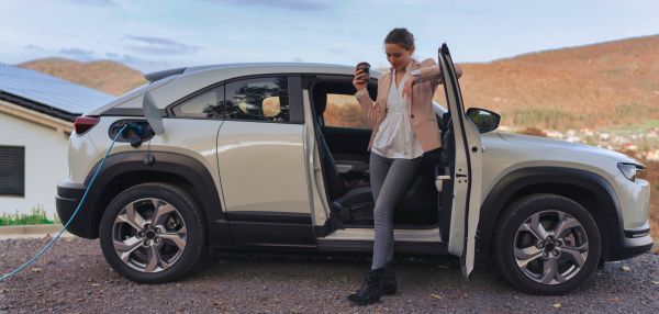 Young woman with smartphone waiting while electric car charging in home charging station, sustainable and economic transportation concept.