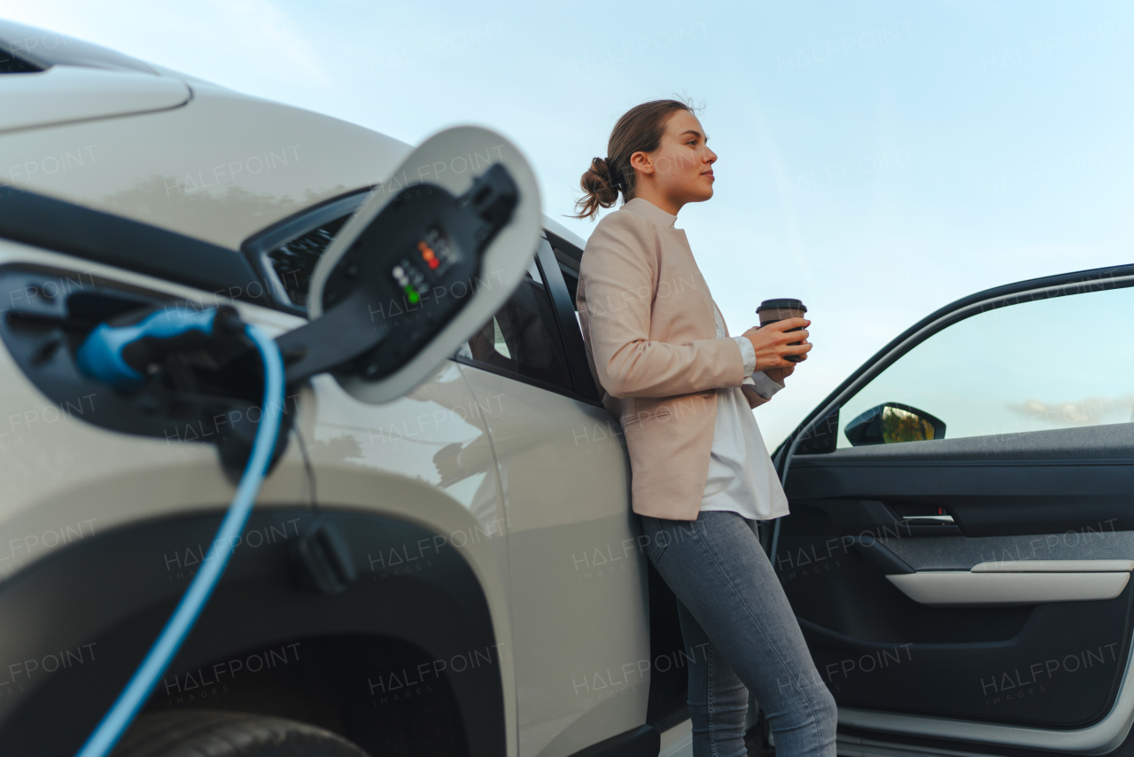 Young woman with cup of coffee waiting while electric car charging, sustainable and economic transportation concept.