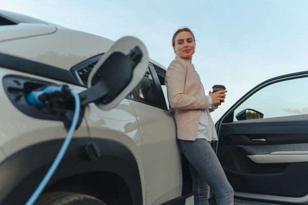 Young woman with cup of coffee waiting while electric car charging, sustainable and economic transportation concept.