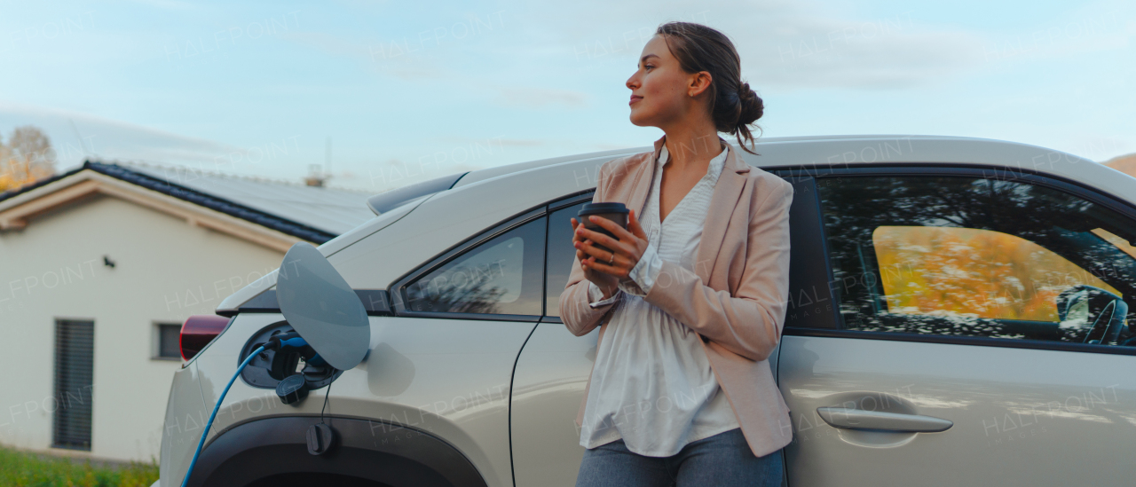 Young woman with cup of coffee waiting while electric car charging, sustainable and economic transportation concept.