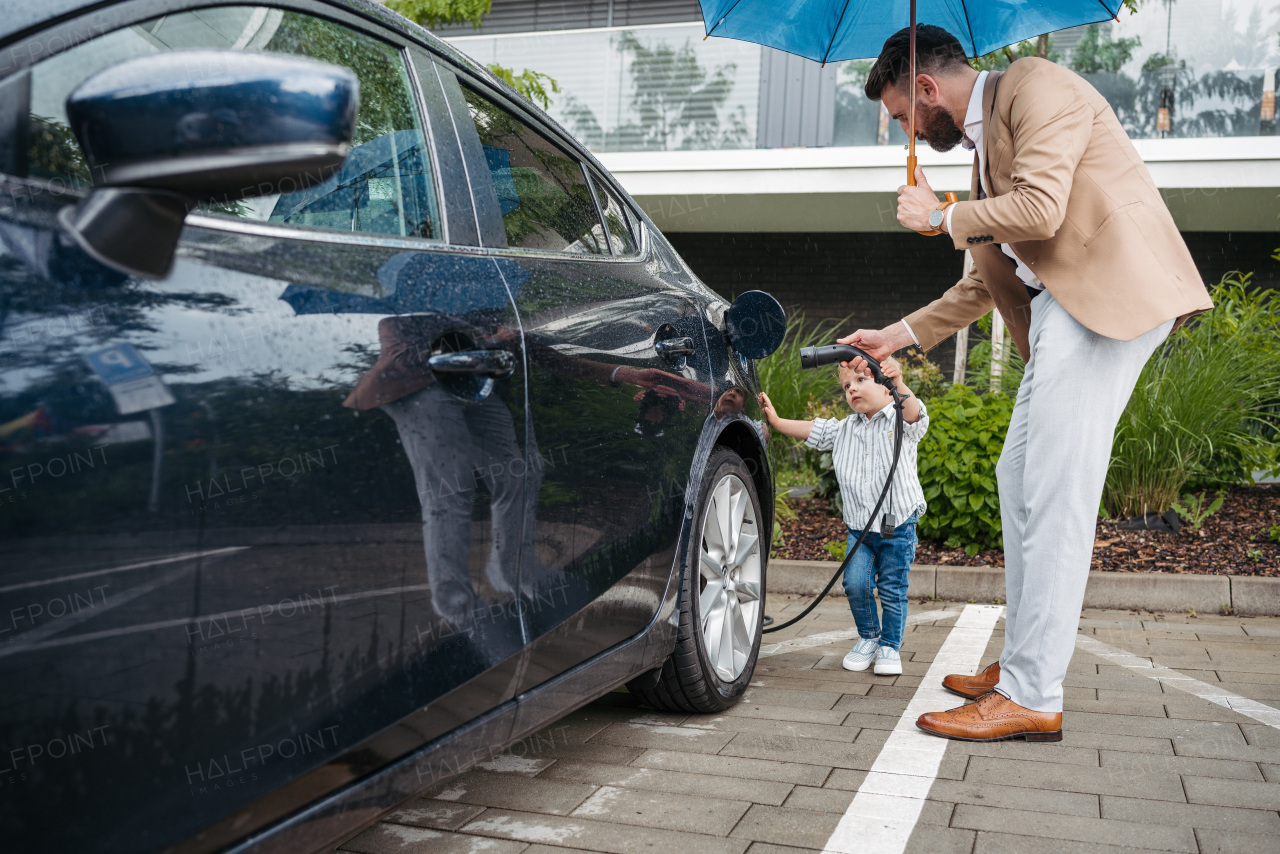 Mature man and his little son charging their electric car on a rainy day, standing under umbrella. Potential risks with charging an electric vehicle on the rain.