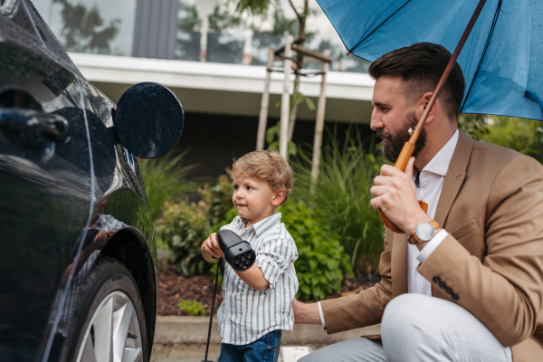 Fathern and little son charging their electric car on a rainy day, standing under umbrella. Potential risks with charging an electric vehicle in the rain. Cute boy helping to plug in charger.