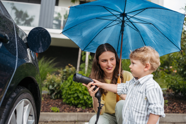 Smiling woman and her little son charging their electric car on a rainy day, standing under umbrella. Potential risks with charging an electric vehicle in the rain. Cute boy helping to plug in charger.